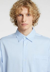 Jasnoniebieska koszula w paski wykonana z tkaniny o miękkiej fakturze. Posiada kołnierzyk z ostrymi końcami, zapięcie na guziki oraz kieszeń na piersi po lewej stronie.