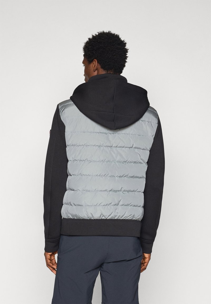 Mężczyzna w czarnej bluzie z kapturem i szarej kamizelce z podszewką, plecami do kamery, na tle białego tła.