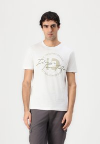 Hvit bomulls T-skjorte med rund hals og korte ermer. Sentral grafikk i beige med tekst "THE ORIGINAL BRAND" og "Denim."