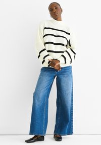 Maglione lavorato a maglia grossa color crema con righe orizzontali nere, abbinato a jeans in denim blu a gamba larga e ballerine nere, su sfondo bianco.