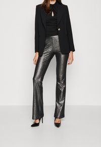 Zwarte blazer met een enkele goudkleurige knoop, een fitted zwarte top met een keyhole-detail en glanzende zilveren flare-broeken; zwarte puntige hakken maken de look af.