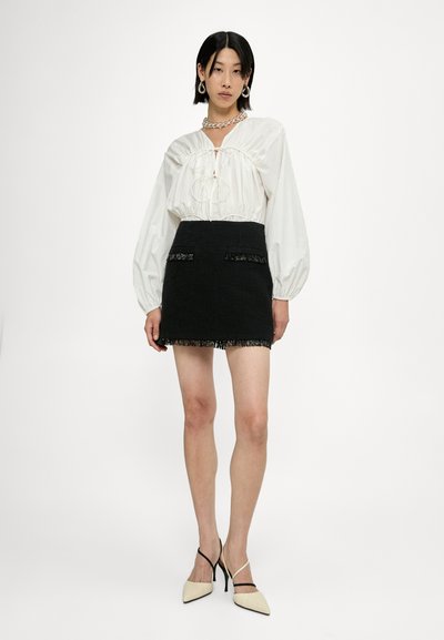 Blouse blanche avec détail froncé, mini jupe en tweed noire avec des franges et des poches, et escarpins beiges à bout pointu avec des brides noires.