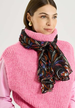 Mujer con pendientes de lágrima dorados, un chaleco de cuello alto rosa de punto y una bufanda negra con estampados de plumas multicolores atada alrededor del cuello.