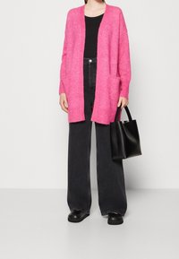 Růžový, oversized cardigan z měkkého materiálu, s otevřeným předním dílem a bočními kapsami, kombinovaný s černým přiléhavým topem a černými džínami s vysokým pasem.