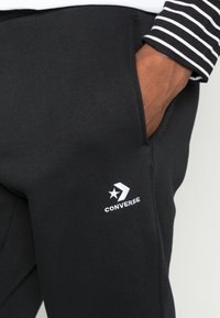 Person som bär svarta Converse-sweatpants med handen i fickan och en svart och vit randig ärm synlig.