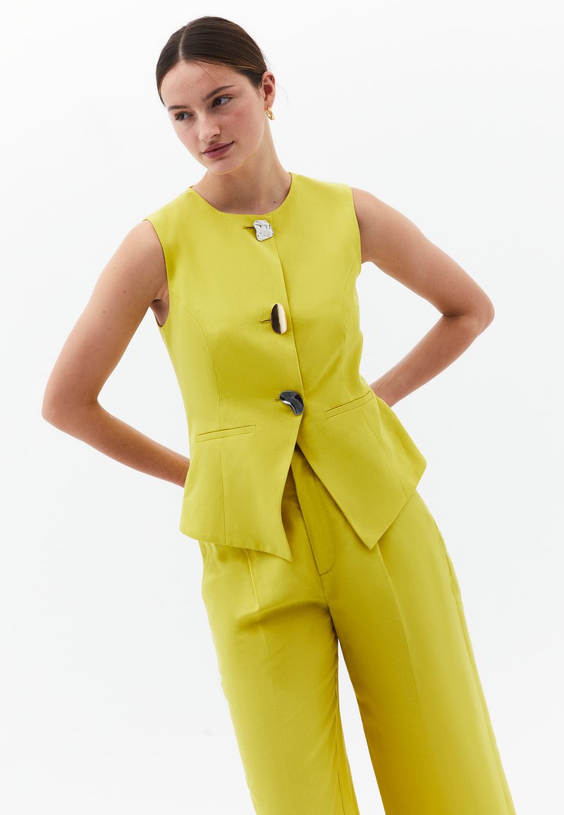 Gilet giallo smanicato con design a peplum, caratterizzato da tre bottoni decorativi di diversi colori. I pantaloni gialli abbinati completano l'outfit.