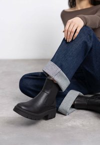 Stivali alla caviglia in pelle nera con pannelli laterali elasticizzati, suole in gomma spessa e dettagli testurizzati, abbinati a jeans scuri in denim con risvolto.