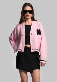 Cardigan rosa a coste con strisce nere, caratterizzato da grandi bottoni e un applicazione a "W". Indossato sopra un crop top nero e una gonna asimmetrica.