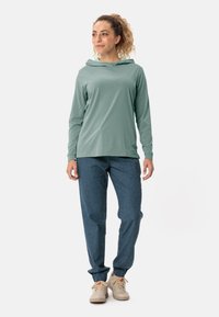 Hellgrüner Hoodie mit langen Ärmeln und entspannter Passform, ausgestattet mit einer Kapuze und einem weichen Stoff. Kombiniert mit dunkelblauen, schmal zulaufenden Hosen und beigen Schuhen.