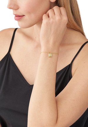 Bracciale d'oro con design a lucchetto, caratterizzato da una delicata catena e un piccolo ciondolo quadrato, indossato al polso di una persona in una maglia nera.