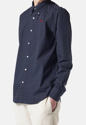 Hombre con camisa azul marino abotonada con pequeño patrón de puntos blancos y logotipo bordado en rojo, combinado con pantalones de pana blancos, mano en el bolsillo.