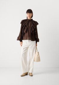 Brun bluse med flæser, høj hals og lange ærmer, parret med hvide vide bukser og beige hælede støvler. Holder en кремfarvet håndtaske.