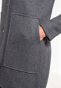 Cappotto di lana grigio con una superficie testurizzata, dotato di una grande tasca applicata e chiusura a bottoni sul davanti. Vestibilità sartoriale con maniche lunghe.