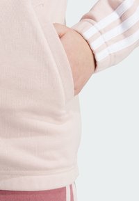 Sudadera de color rosa claro con textura acanalada, bolsillo lateral y rayas blancas en la manga. Acabado de tejido suave. Diseño ceñido.