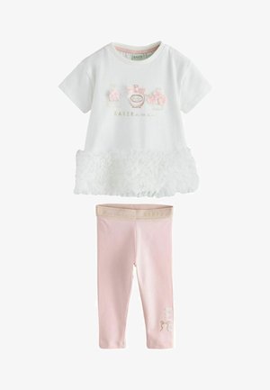 Ung pige med kort sort hår iført en hvid kjole med blomsterdetaljer, pink leggings og hvide sneakers, stående på en grå gulv.