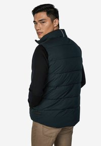 Dunkelgrüne Steppweste mit horizontalem Steppmuster, hohem Kragen und Seitentasche, getragen über einem schwarzen Langarmshirt. Verstellbare Passform.