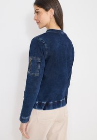 Veste en denim bleu foncé avec une poche zippée sur la manche, des poignets et un ourlet côtelés. Présente une coupe décontractée et une texture douce, idéale pour superposer.