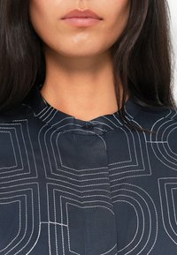 Camicia blu navy con tessuto testurizzato, dotata di dettagli con bottoni sul colletto e cuciture geometriche bianche lungo la parte anteriore.