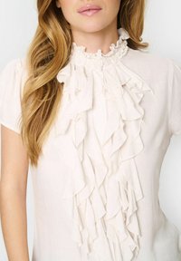 Lichtroze blouse met een gerimpelde voorkant, hoge gerimpelde kraag, korte mouwen en knoopsluiting. Gemaakt van lichte materialen.