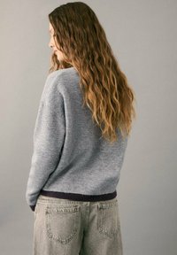 Svijetlosivi pulover širokog kroja s tamnim rebrastim rubom, uparen s visokim sivim trapericama. Kosa u labavim valovima.