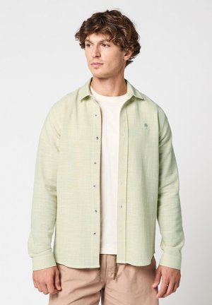 Junger Mann mit lockigem Haar, der ein hellgrünes Hemd über einem weißen T-Shirt und beige Hose trägt und zur Seite blickt.