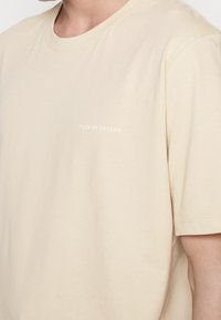 Närbild av en person som bär en beige T-shirt med kort ärm där det står "TIGER OF SWEDEN" tryckt med små vita bokstäver på bröstet.