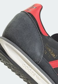 Svart mocka- och tygsneaker med röda accenter, texturerad profil, vit mellansula och svart gummisul med greppmönster.