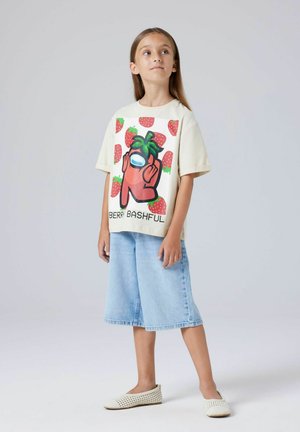 Jeune fille debout portant un T-shirt crème avec un personnage fraise rouge et le texte "BERRY BASHFUL", un short en jean clair et des chaussures blanches sans lacets.