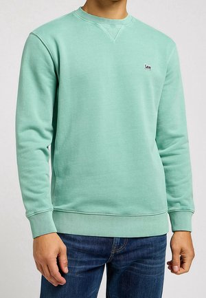 Mężczyzna ubrany w miętowo-zielony sweter crewneck z małym logo Lee na piersi oraz niebieskie dżinsy, stojący na neutralnym tle.