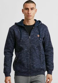 Sweat à capuche bleu marine avec un motif texturé, fermeture éclair, poches devant et une capuche intérieure contrastante. Agrémenté d'un petit patch logo.