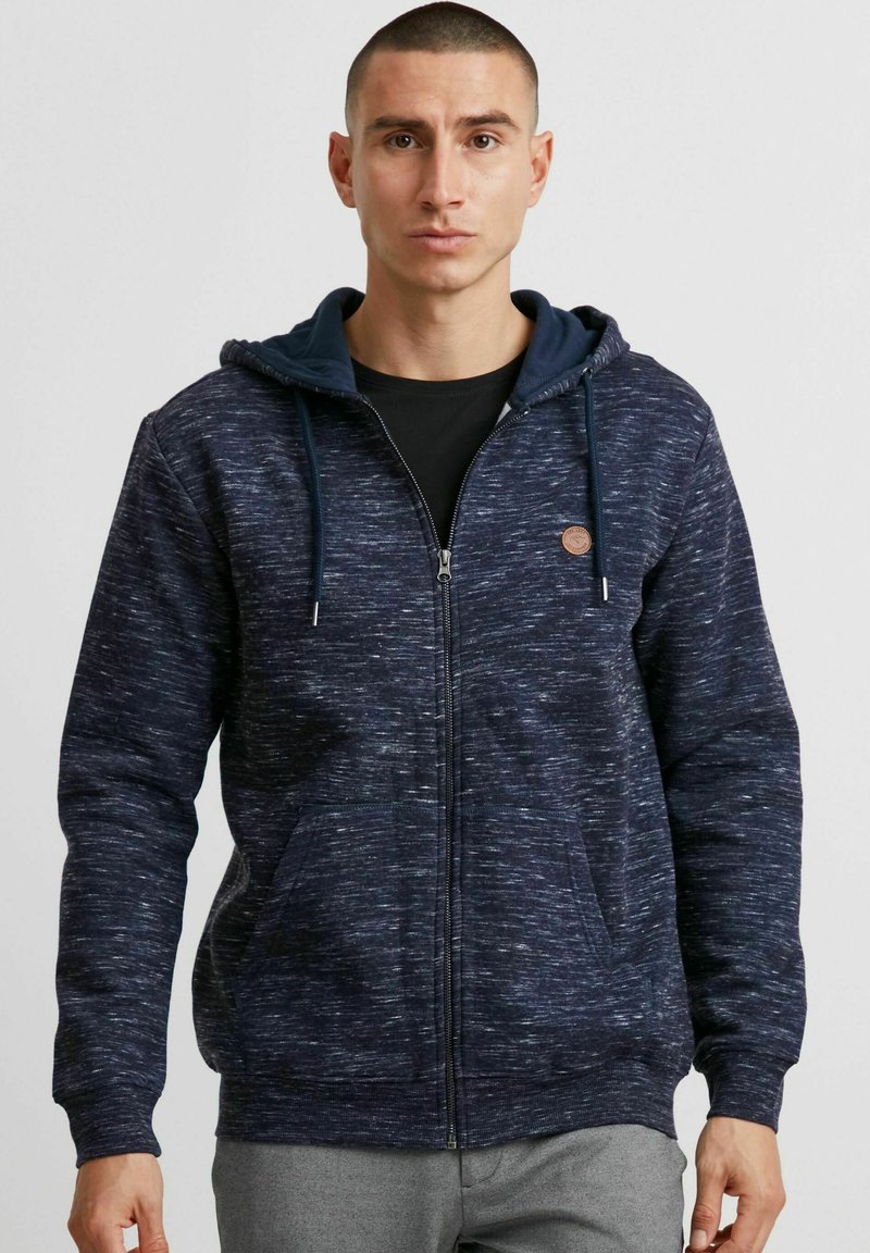 Sweat à capuche bleu marine avec un motif texturé, fermeture éclair, poches devant et une capuche intérieure contrastante. Agrémenté d'un petit patch logo.