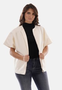 Giacca bianca oversize con zip frontale, maniche corte, tessuto strutturato e vestibilità rilassata, abbinata a un dolcevita nero e jeans.