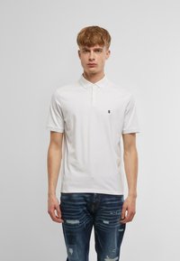 Balta polo krekls ar apkakli un trim pogu slēdzeni, ar mazu logotipu uz kreisā krūts. Valkāts ar nolietotām zilām džinsiem.