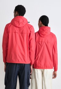 Chaquetas ligeras rojas con capuchas y puños elásticos. Presentan un cordón y costuras visibles. Llevadas sobre pantalones azul marino y crema.