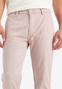 Ljusrosa bomullsshorts med en slät textur, dragkedja, enkel knäppning och bälteshällor. Har minimala sömdetaljer.