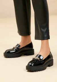 Fekete lakkbőr mokaszinek vastag platformtalppal és díszes aranylánc részlettel az elején. Simás, fényes kivitel.