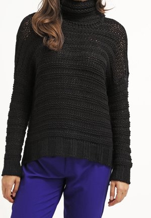 Black knitted sweater with a high neckline, ribbed detailing, and loose fit. Long sleeves with open weave sections. Paired with blue trousers.