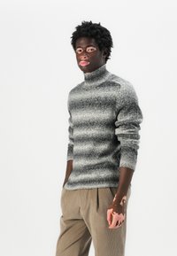 Pull en col roulé dégradé gris et noir en tissu tricoté texturé, avec des manches longues et des poignets côtelés. Associé à un pantalon clair.