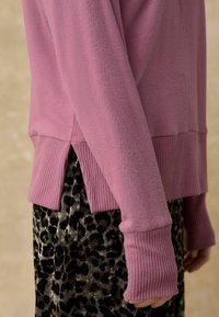 Różowy sweter z dzianiny z prążkowanymi detalami na dole i mankietach, z bocznymi rozcięciami. W połączeniu z spódnicą w panterkę i teksturowanym materiałem.