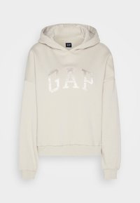 Cremfarbener, cropped Hoodie aus weichem Stoff mit einer Kapuze und glänzendem "GAP"-Logo auf der Vorderseite.