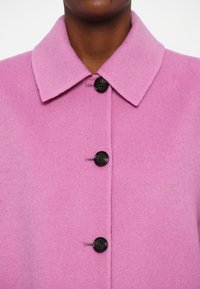 Manteau en laine rose avec un col à revers, présentant quatre boutons noirs sur le devant. La texture semble lisse et légèrement duveteuse.