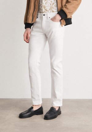 Personne portant un pantalon blanc slim, des mocassins noirs, une chemise à motifs et une veste marron avec une main dans la poche, debout sur un sol gris.