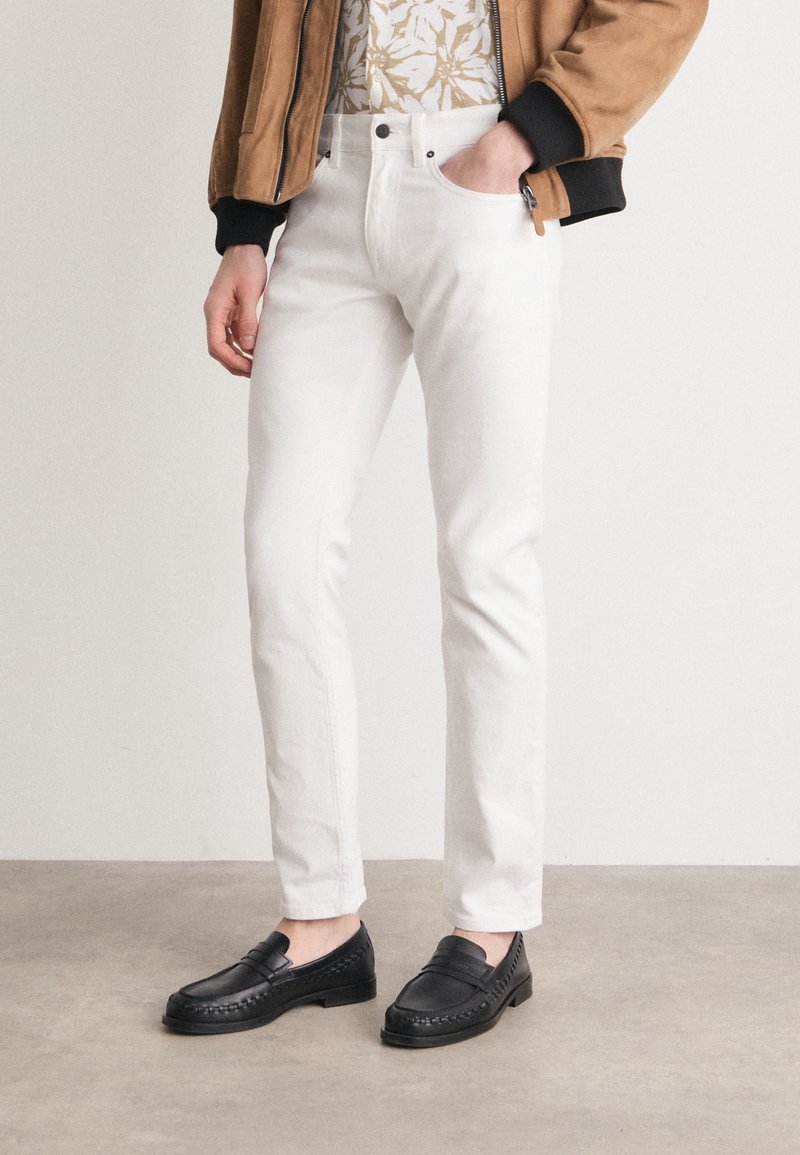 Personne portant un pantalon blanc slim, des mocassins noirs, une chemise à motifs et une veste marron avec une main dans la poche, debout sur un sol gris.