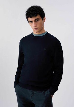 Giovane uomo con capelli ricci e scuri che indossa un maglione nero girocollo sopra una camicia blu, in piedi con le mani nelle tasche davanti a uno sfondo semplice.