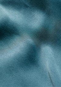 Tissu bleu-vert à la texture lisse légèrement froissée, montrant des fils finement tissés et des reflets doux de lumière.