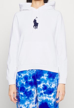 Person iført en hvid hættetrøje med sort polo spiller-logo på brystet og blå tie-dye bukser, stående mod en ensfarvet baggrund.