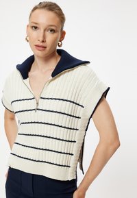Stickad tröja med marinblå krage, dragkedja, korta ärmar, krämfärgad bas med marinblå ränder, med ribbad textur och sidoflikar.