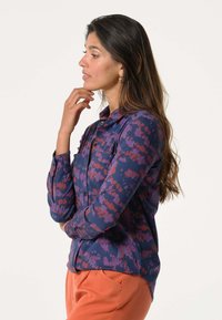 Een overhemd met knopen in een donkerblauwe stof met rode en paarse abstracte patronen. Voorzien van een kraag, een borstzak en lange mouwen.