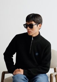 Maglione a coste nero con colletto alto e zip, caratterizzato da un logo blu; abbinato a grandi occhiali da sole quadrati neri. Jeans casual in denim.