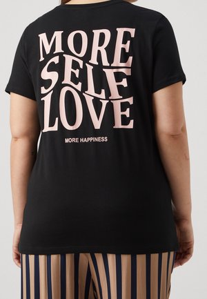 Osoba nosząca czarną koszulkę z różowym napisem "WIĘCEJ MIŁOŚCI DO SIEBIE" oraz "WIĘCEJ SZCZĘŚCIA" w połączeniu z beżowymi i granatowo paskowanymi spodniami.